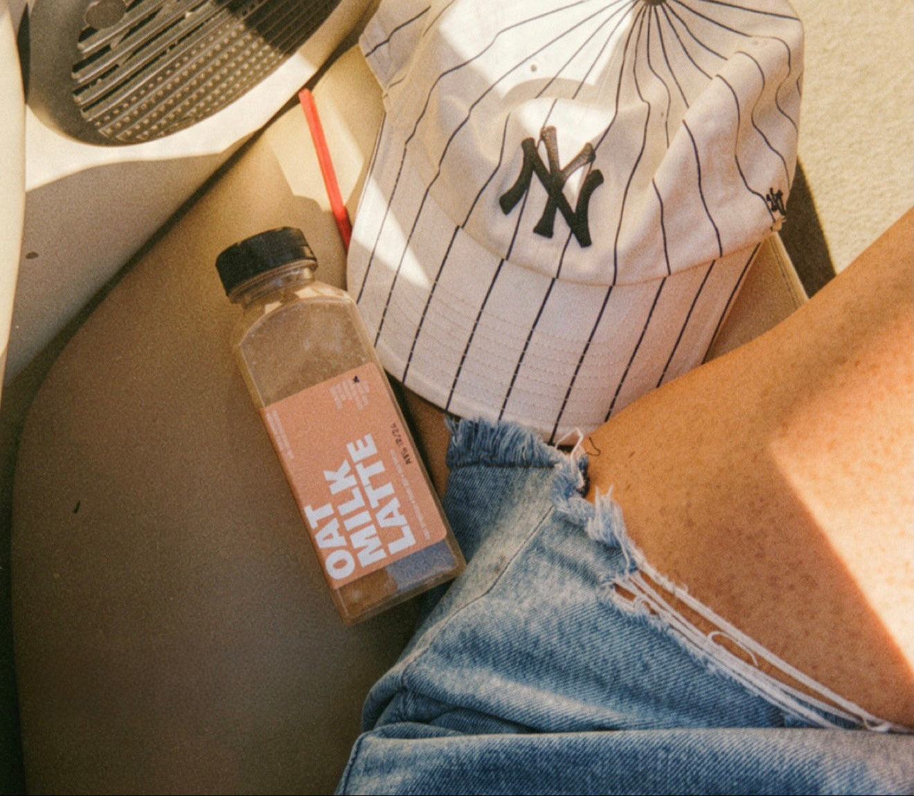 A bottle of Corso Coffee Oat Milk Latte resting on a beige boat seat next to a person's denim shorts and baseball cap
